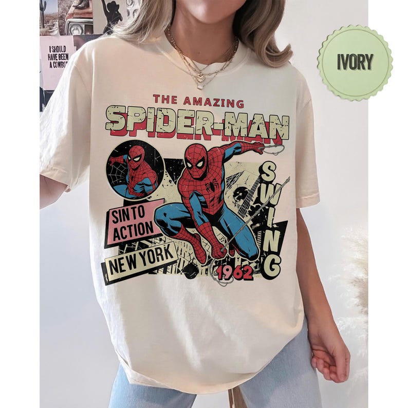 Op de afbeelding: Ivoor kleurig T-shirt met een vintage Spider-Man graphic. Het ontwerp bevat de tekst "THE AMAZING SPIDER-MAN", "SIN TO ACTION" en "NEW YORK", samen met een illustratie van Spider-Man. Het shirt heeft een retro stripboek stijl.