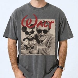May include: Dark gray t-shirt featuring a vintage image of Walt Disney and Mickey Mouse wearing sunglasses. The word "Walt" is printed in red above the image. The shirt has a classic crew neck and short sleeves.