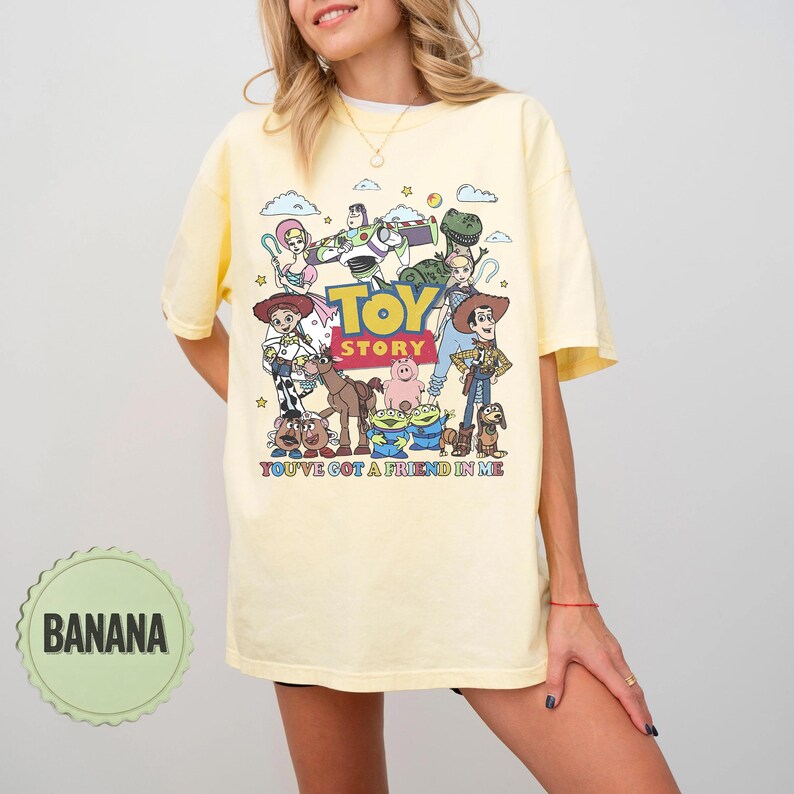 May include: A light yellow t-shirt featuring a colorful graphic of characters from the animated film "Toy Story." The design includes the film's title and the phrase "You've Got a Friend in Me."