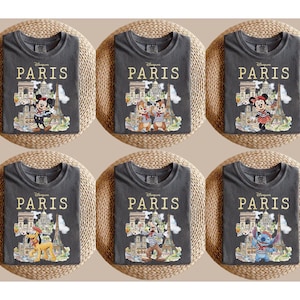 K&ouml;nnte beinhalten: Sechs anthrazitfarbene T-Shirts mit Disney-Figuren in Paris. Jedes Shirt hat den Schriftzug "PARIS" in Gold &uuml;ber einer Illustration des Eiffelturms und anderer Wahrzeichen. Zu den Figuren geh&ouml;ren Micky Maus, Minnie Maus, Chip und Chap, Pluto und Stitch.