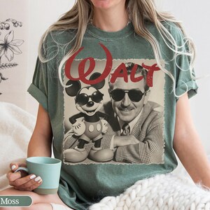 May include: Moss green t-shirt featuring a vintage-style graphic of Mickey Mouse wearing sunglasses and Walt Disney. The word "WALT" is printed in red above the image. The shirt is a relaxed fit.