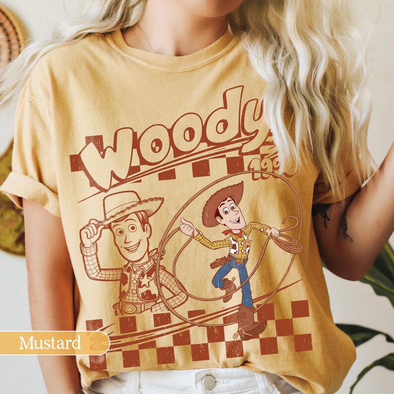 Puede incluir: Camiseta de color mostaza con un dise&ntilde;o de Woody de Toy Story. El dise&ntilde;o incluye la palabra "Woody" sobre dos ilustraciones del personaje, con un fondo a cuadros. Prenda de vestir informal.