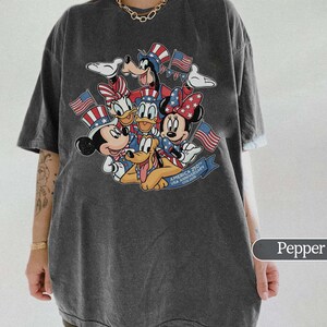 May include: Pepper-colored t-shirt featuring a graphic of Mickey, Minnie, Donald, Daisy, Goofy, and Pluto, all in patriotic attire with American flags. The graphic includes the text "AMERICA 250th USA ANNIVERSARY 1776-2026".
