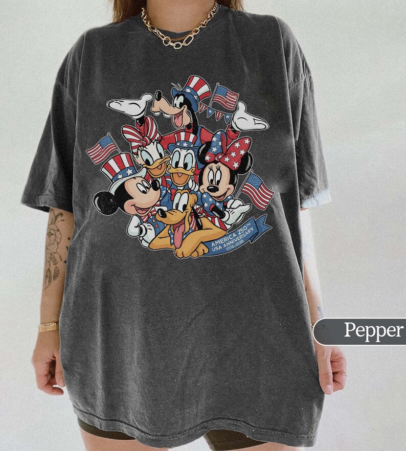 May include: Pepper-colored t-shirt featuring a graphic of Mickey, Minnie, Donald, Daisy, Goofy, and Pluto, all in patriotic attire with American flags. The graphic includes the text "AMERICA 250th USA ANNIVERSARY 1776-2026".
