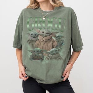 Op de afbeelding: Olijfgroen T-shirt met een afbeelding van Grogu uit The Mandalorian. Het ontwerp bevat meerdere Grogu-afbeeldingen en het woord "GROGU" in een groene, reli&euml;fletter. Het T-shirt heeft een relaxte pasvorm.