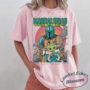 May include: A light pink short-sleeve t-shirt featuring a graphic print of The Mandalorian and Grogu. The text reads THE MANDALORIAN at the top and THIS IS THE WAY at the bottom. The design includes a red sun, grey clouds, and teal accents.