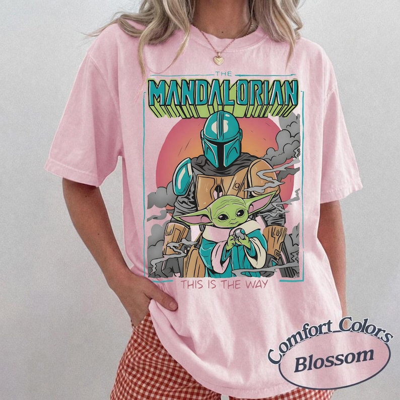 May include: A light pink short-sleeve t-shirt featuring a graphic print of The Mandalorian and Grogu. The text reads THE MANDALORIAN at the top and THIS IS THE WAY at the bottom. The design includes a red sun, grey clouds, and teal accents.