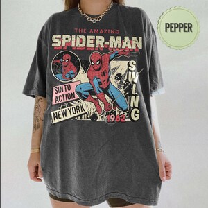 Op de afbeelding: Donkergrijs t-shirt met een vintage Spider-Man graphic. Het ontwerp bevat de woorden "The Amazing Spider-Man", "SINTO ACTION NEW YORK" en "1962". De superheld wordt afgebeeld in een dynamische pose, met rode en blauwe kleuren.