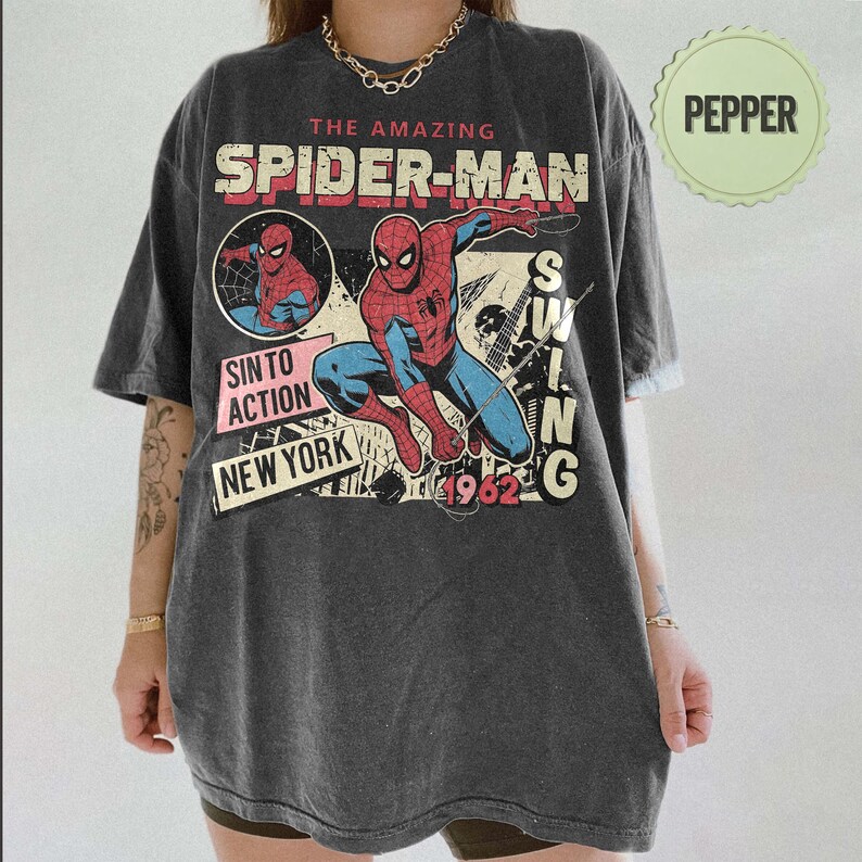 Op de afbeelding: Donkergrijs t-shirt met een vintage Spider-Man graphic. Het ontwerp bevat de woorden "The Amazing Spider-Man", "SINTO ACTION NEW YORK" en "1962". De superheld wordt afgebeeld in een dynamische pose, met rode en blauwe kleuren.