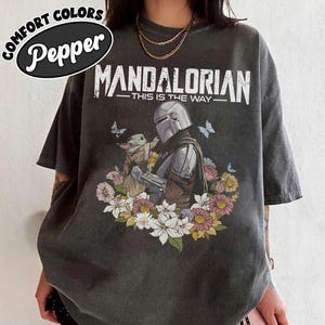 May include: Dark grey t-shirt featuring a Mandalorian graphic with Grogu surrounded by flowers and butterflies. The text "MANDALORIAN - THIS IS THE WAY" is printed above the image. The shirt has the text "COMFORT COLORS Pepper" in a circular design.