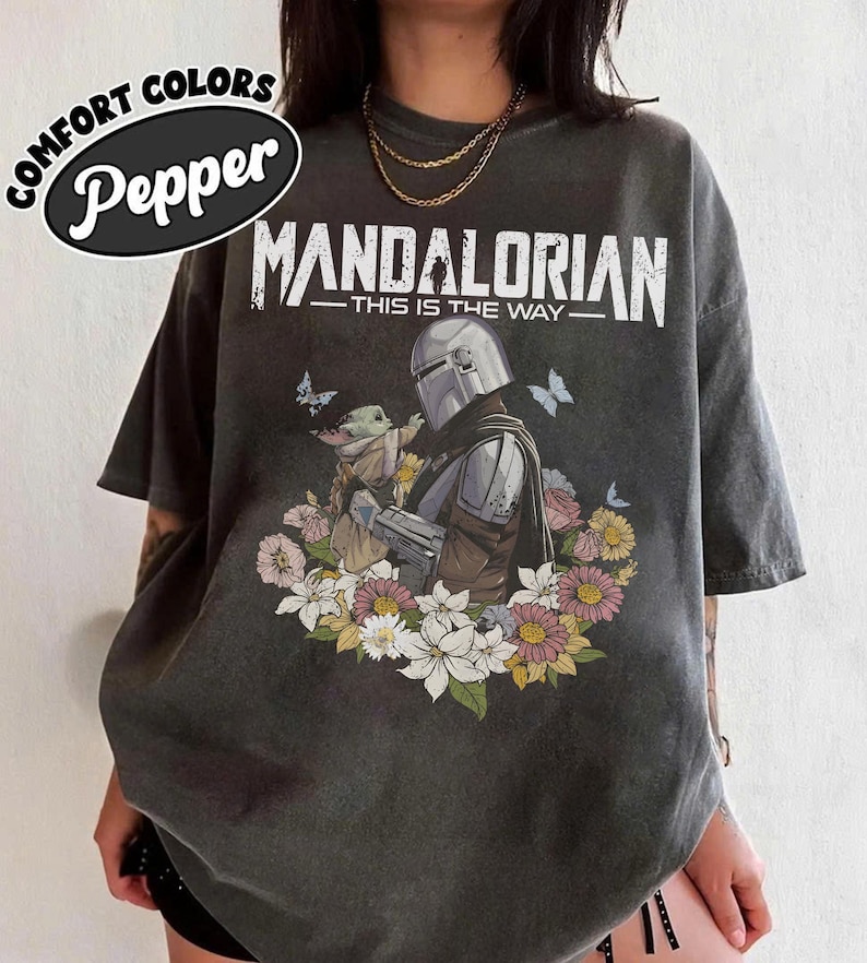 May include: Dark grey t-shirt featuring a Mandalorian graphic with Grogu surrounded by flowers and butterflies. The text "MANDALORIAN - THIS IS THE WAY" is printed above the image. The shirt has the text "COMFORT COLORS Pepper" in a circular design.