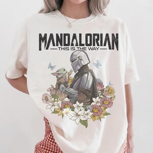 May include: Cream-colored t-shirt featuring a graphic of The Mandalorian and Grogu surrounded by flowers and butterflies. The text "MANDALORIAN - THIS IS THE WAY" is printed across the top in black.