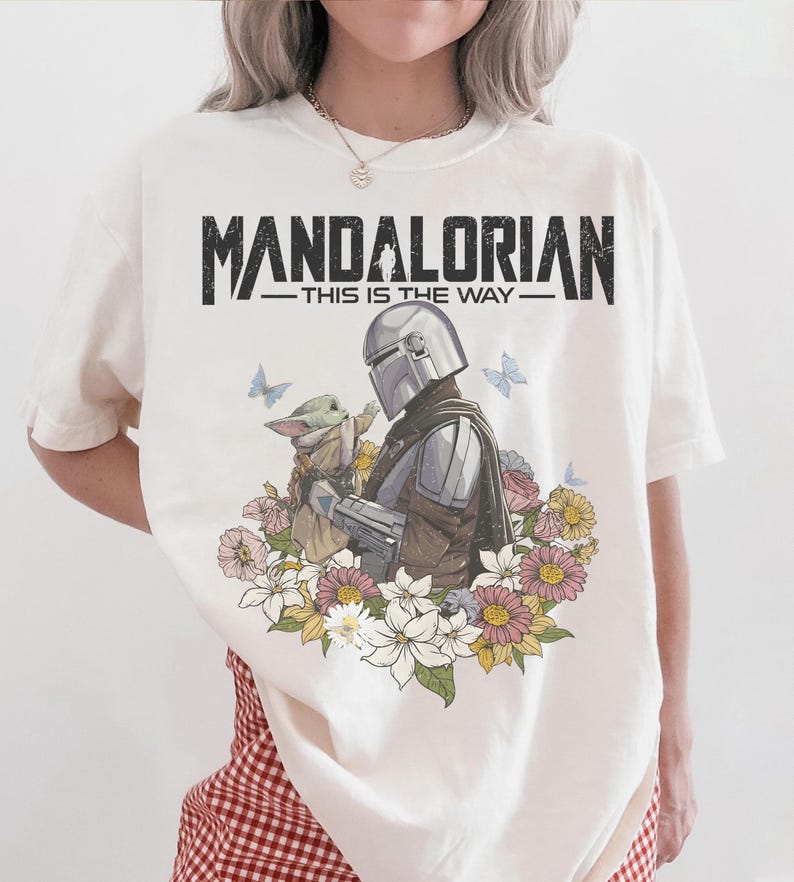 May include: Cream-colored t-shirt featuring a graphic of The Mandalorian and Grogu surrounded by flowers and butterflies. The text "MANDALORIAN - THIS IS THE WAY" is printed across the top in black.