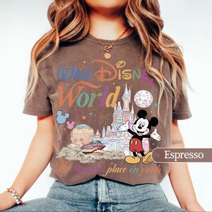 May include: Espresso-colored t-shirt featuring a colorful graphic of Mickey Mouse in front of a castle, with the words "Walt Disney World" and "The happiest place on earth". The design includes balloons, fireworks, and a planet.