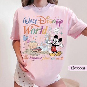 May include: A light pink t-shirt featuring a colorful graphic of Mickey Mouse with the words "Walt Disney World" and "The happiest place on earth". The design includes a castle, fireworks, and balloons.