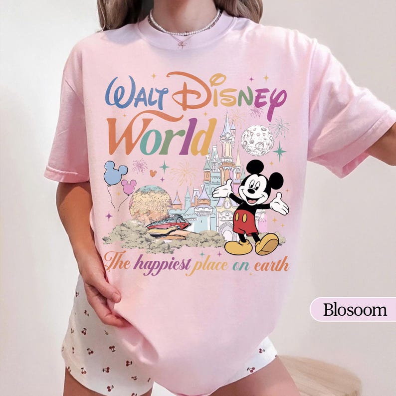 May include: A light pink t-shirt featuring a colorful graphic of Mickey Mouse with the words "Walt Disney World" and "The happiest place on earth". The design includes a castle, fireworks, and balloons.