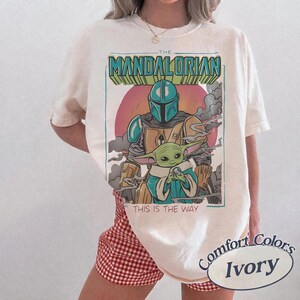 May include: An ivory short-sleeve t-shirt featuring a graphic print of The Mandalorian and Grogu. The design includes the text THE MANDALORIAN at the top and THIS IS THE WAY at the bottom, set against a circular sunset background with smoke elements.