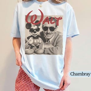 May include: Light blue t-shirt featuring a vintage-style graphic of Walt Disney and Mickey Mouse wearing sunglasses. The word "Walt" is in red script above the image. The shirt is a chambray color.
