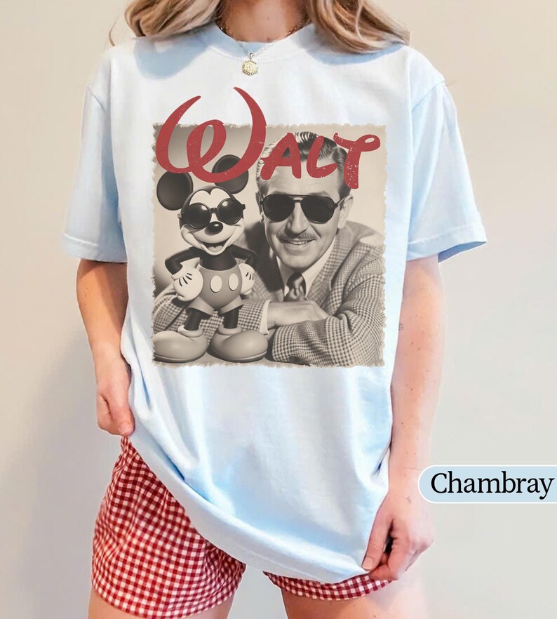 May include: Light blue t-shirt featuring a vintage-style graphic of Walt Disney and Mickey Mouse wearing sunglasses. The word "Walt" is in red script above the image. The shirt is a chambray color.