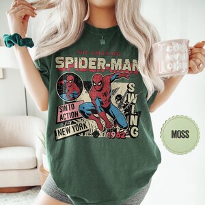 Op de afbeelding: Mosgroen t-shirt met een vintage Spider-Man graphic. Het ontwerp bevat de tekst "THE AMAZING SPIDER-MAN", "SIN TO ACTION", "NEW YORK" en "1962". De graphic toont Spider-Man in actie in een retro stripboekstijl.
