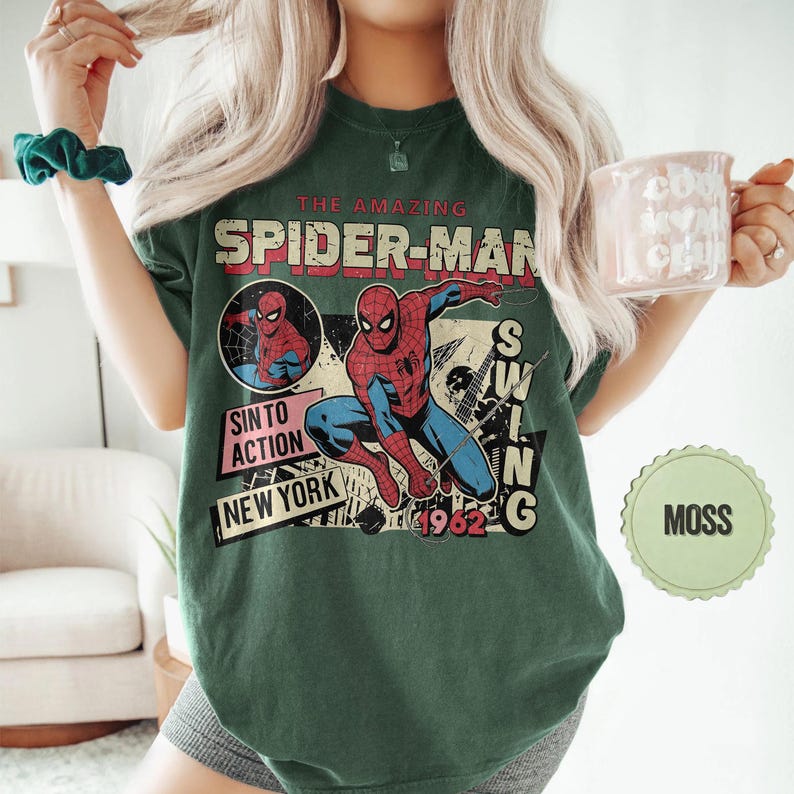 Op de afbeelding: Mosgroen t-shirt met een vintage Spider-Man graphic. Het ontwerp bevat de tekst "THE AMAZING SPIDER-MAN", "SIN TO ACTION", "NEW YORK" en "1962". De graphic toont Spider-Man in actie in een retro stripboekstijl.