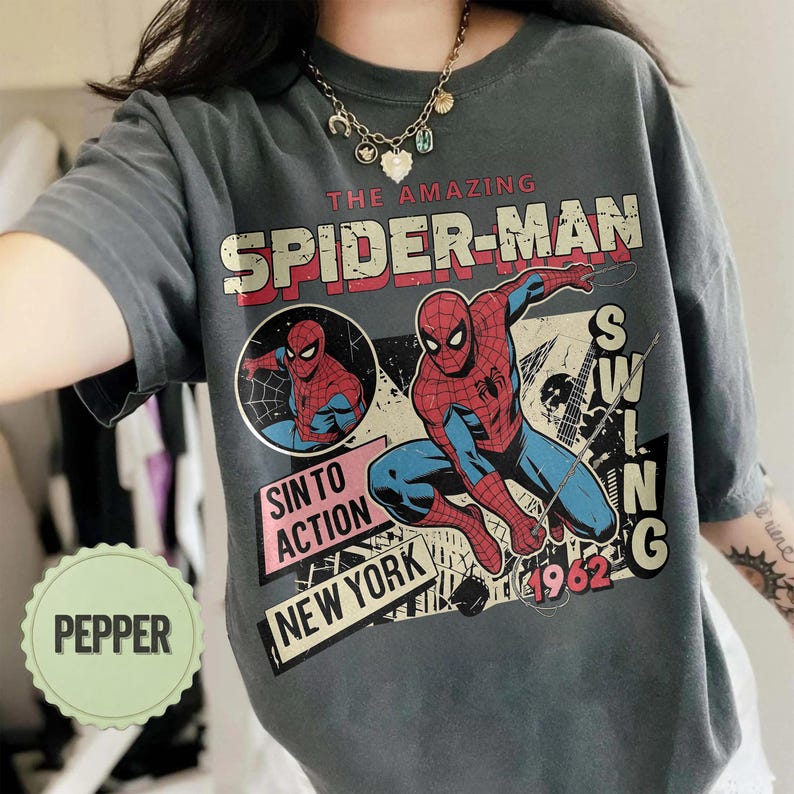 Op de afbeelding: Donkergrijs T-shirt met een vintage Spider-Man-afbeelding. Het ontwerp toont Spider-Man in actie, met de tekst "THE AMAZING SPIDER-MAN" in rood en wit. Extra tekst omvat "SIN TO ACTION" en "NEW YORK".