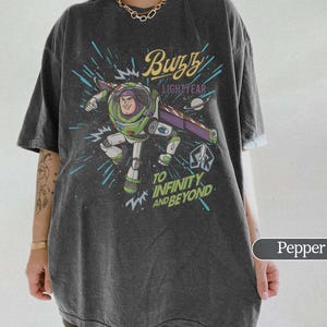 Puede incluir: Camiseta color pimienta con un dise&ntilde;o de Buzz Lightyear de Toy Story. El dise&ntilde;o incluye el texto "Buzz Lightyear" y "To Infinity and Beyond" sobre un fondo espacial. La camiseta tiene mangas cortas y un corte holgado.
