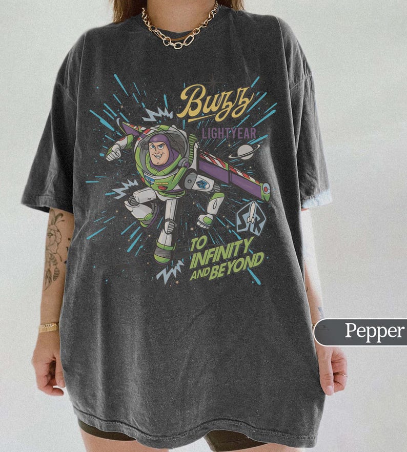 Puede incluir: Camiseta color pimienta con un dise&ntilde;o de Buzz Lightyear de Toy Story. El dise&ntilde;o incluye el texto "Buzz Lightyear" y "To Infinity and Beyond" sobre un fondo espacial. La camiseta tiene mangas cortas y un corte holgado.