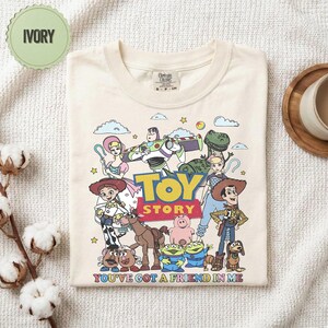 May include: Ivory-colored t-shirt featuring a colorful graphic of characters from the animated film "Toy Story." The design includes Woody, Buzz Lightyear, and other beloved characters, with the text "TOY STORY" and "YOU'VE GOT A FRIEND IN ME."