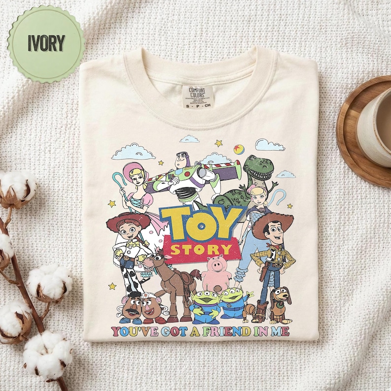 May include: Ivory-colored t-shirt featuring a colorful graphic of characters from the animated film "Toy Story." The design includes Woody, Buzz Lightyear, and other beloved characters, with the text "TOY STORY" and "YOU'VE GOT A FRIEND IN ME."
