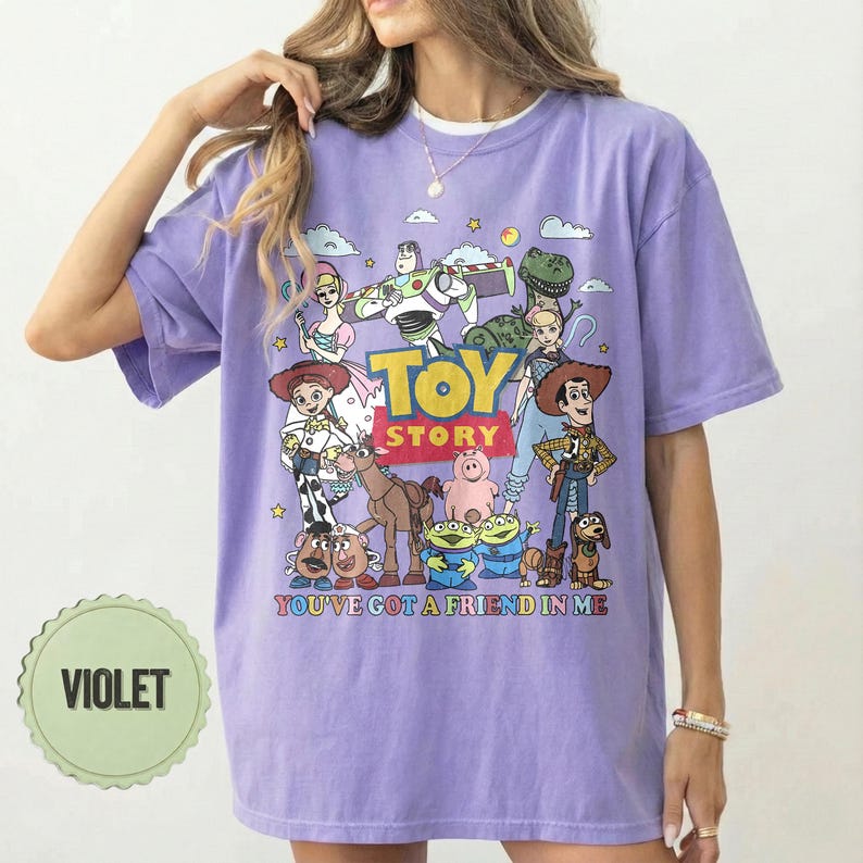 May include: Violet t-shirt featuring a colorful graphic of Toy Story characters. The design includes Woody, Buzz Lightyear, and other characters from the movie, with the text "TOY STORY" and "YOU'VE GOT A FRIEND IN ME".