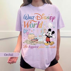 May include: Orchid-colored t-shirt featuring a colorful graphic of Mickey Mouse, a castle, and the words "Walt Disney World" and "The happiest place on earth." The design includes fireworks, balloons, and a planet.