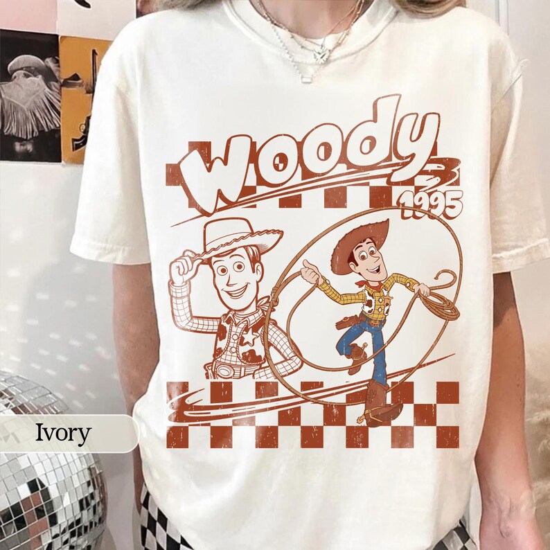 Puede incluir: Camiseta color marfil con un gr&aacute;fico marr&oacute;n del personaje Woody de la pel&iacute;cula Toy Story. El dise&ntilde;o incluye la palabra "Woody" y el a&ntilde;o "1995". La camiseta tiene un patr&oacute;n de tablero de ajedrez marr&oacute;n y blanco.