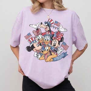 May include: A light purple t-shirt featuring a graphic of Disney characters Mickey Mouse, Minnie Mouse, Donald Duck, Daisy Duck, Goofy, and Pluto, all dressed in patriotic attire and holding American flags. The shirt has the text "AMERICA 250TH USA ANNIVERSARY 1776-2026".