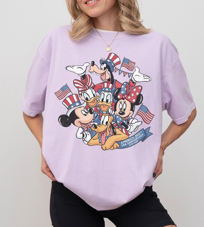 May include: A light purple t-shirt featuring a graphic of Disney characters Mickey Mouse, Minnie Mouse, Donald Duck, Daisy Duck, Goofy, and Pluto, all dressed in patriotic attire and holding American flags. The shirt has the text "AMERICA 250TH USA ANNIVERSARY 1776-2026".