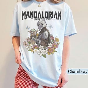 May include: Light blue t-shirt featuring the text "MANDALORIAN" above an illustration of the Mandalorian and Grogu surrounded by flowers. The phrase "THIS IS THE WAY" is printed below the title. The shirt is a light blue color.