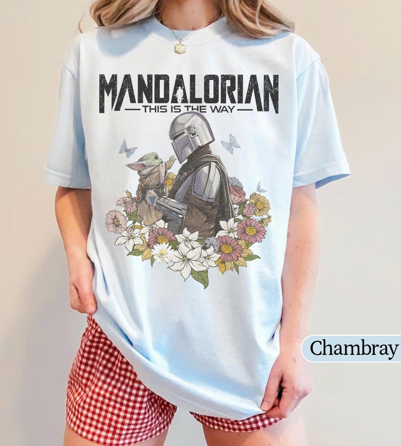 May include: Light blue t-shirt featuring the text "MANDALORIAN" above an illustration of the Mandalorian and Grogu surrounded by flowers. The phrase "THIS IS THE WAY" is printed below the title. The shirt is a light blue color.