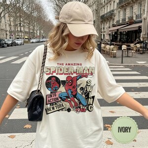 Op de afbeelding: Ivoor T-shirt met een vintage Spider-Man graphic. Het ontwerp bevat de tekst "THE AMAZING SPIDER-MAN", een stripachtige illustratie van Spider-Man en de woorden "SINTO ACTION NEW YORK 1962". Het shirt wordt gedragen met een beige baseball cap.