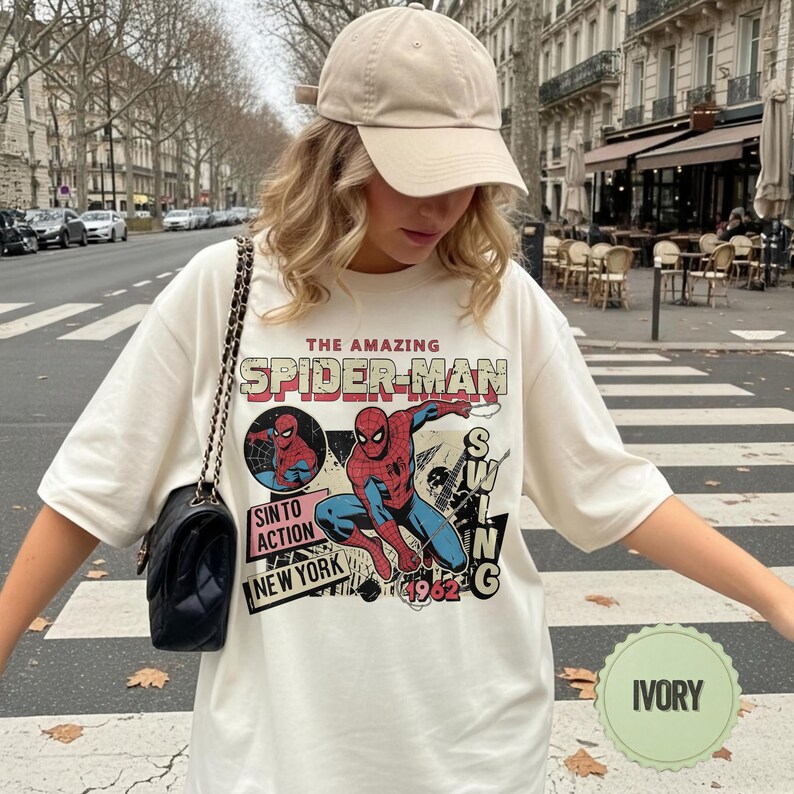 Op de afbeelding: Ivoor T-shirt met een vintage Spider-Man graphic. Het ontwerp bevat de tekst "THE AMAZING SPIDER-MAN", een stripachtige illustratie van Spider-Man en de woorden "SINTO ACTION NEW YORK 1962". Het shirt wordt gedragen met een beige baseball cap.