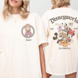 Op de afbeelding: Cr&egrave;mekleurig T-shirt met een Disney World-ontwerp. De voorkant toont een stripfiguur in een geruite cirkel met het woord "Name" eronder. De achterkant heeft een Disney World-afbeelding met personages en de tekst "Est 1971 Florida."