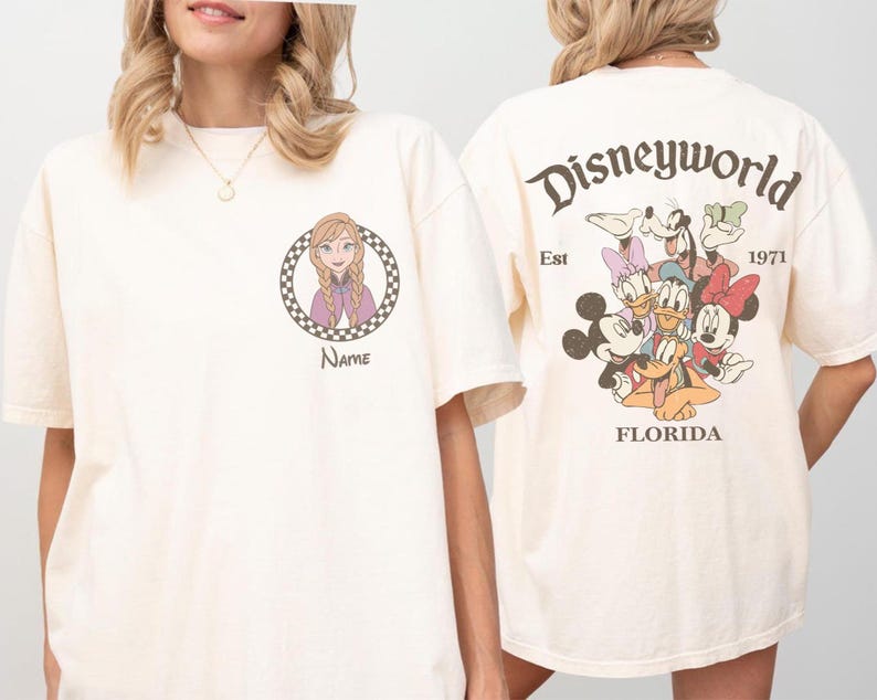 Op de afbeelding: Cr&egrave;mekleurig T-shirt met een Disney World-ontwerp. De voorkant toont een stripfiguur in een geruite cirkel met het woord "Name" eronder. De achterkant heeft een Disney World-afbeelding met personages en de tekst "Est 1971 Florida."