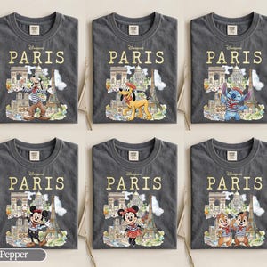 K&ouml;nnte beinhalten: Sechs graue T-Shirts mit dem Wort "PARIS" in goldfarbenen Buchstaben. Jedes Shirt zeigt eine andere Disney-Figur vor Pariser Wahrzeichen wie dem Eiffelturm und dem Arc de Triomphe. Zu den Figuren geh&ouml;ren Goofy, Pluto, Stitch, Mickey Mouse, Minnie Mouse und Chip und Chap.