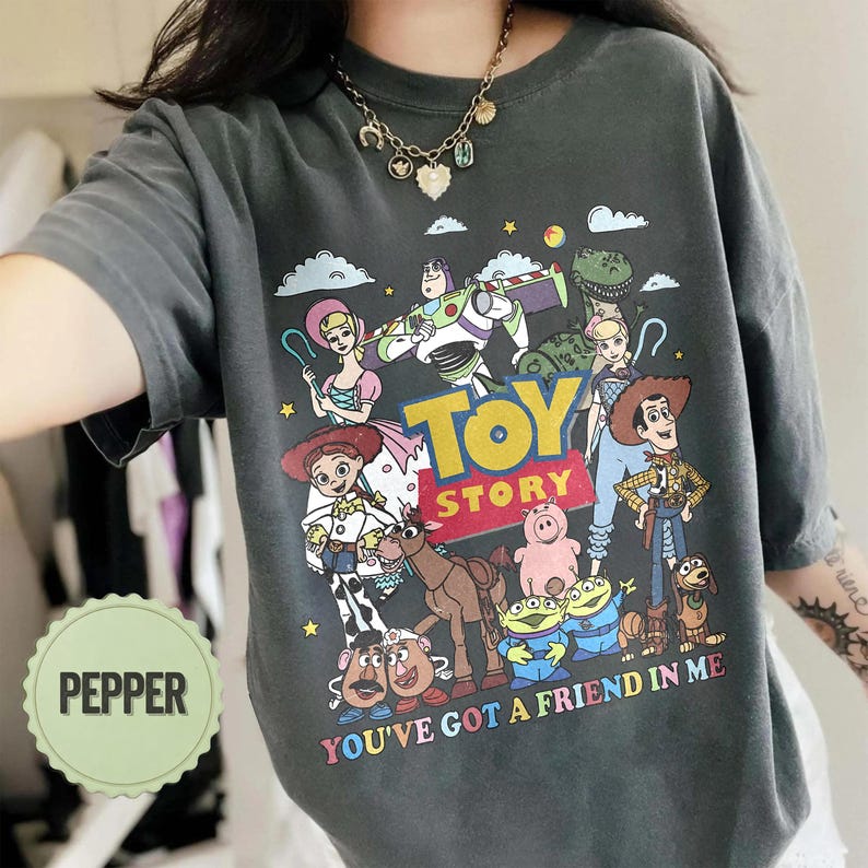 May include: Dark gray t-shirt featuring a colorful graphic of characters from the animated film "Toy Story." The design includes the words "Toy Story" and "You've Got a Friend in Me."