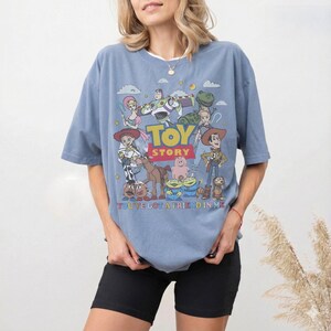 May include: A light blue t-shirt featuring a graphic of characters from the animated film "Toy Story." The design includes Woody, Buzz Lightyear, and other characters, with the words "Toy Story" and "You've Got a Friend in Me."