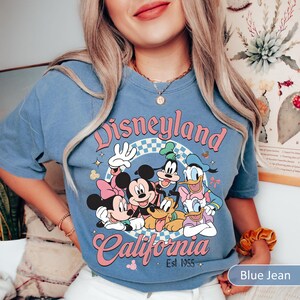May include: Blue jean colored t-shirt featuring a Disneyland California graphic with Mickey Mouse, Minnie Mouse, Goofy, Donald Duck, and Pluto. The graphic is in pink, white, and blue. The text "Disneyland California Est 1955" is also on the shirt.