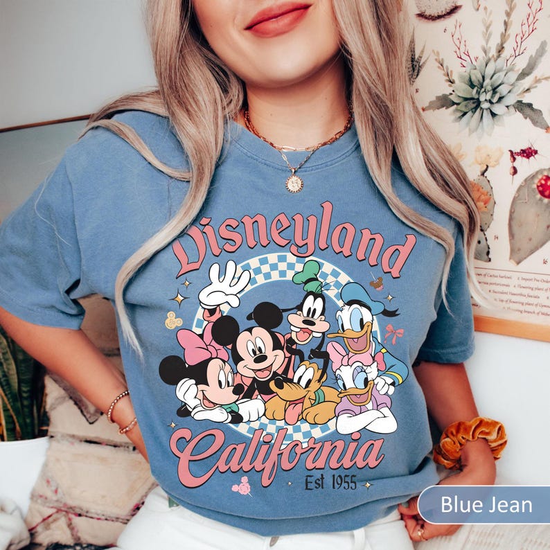 May include: Blue jean colored t-shirt featuring a Disneyland California graphic with Mickey Mouse, Minnie Mouse, Goofy, Donald Duck, and Pluto. The graphic is in pink, white, and blue. The text "Disneyland California Est 1955" is also on the shirt.