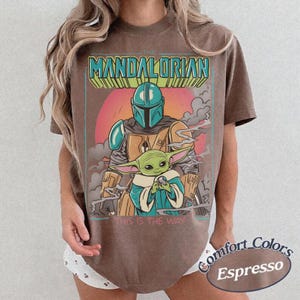 May include: A brown t-shirt featuring a graphic print of The Mandalorian and Grogu. The text THE MANDALORIAN appears at the top in teal and yellow, with THIS IS THE WAY printed at the bottom. The design includes a pink sun and grey smoke.