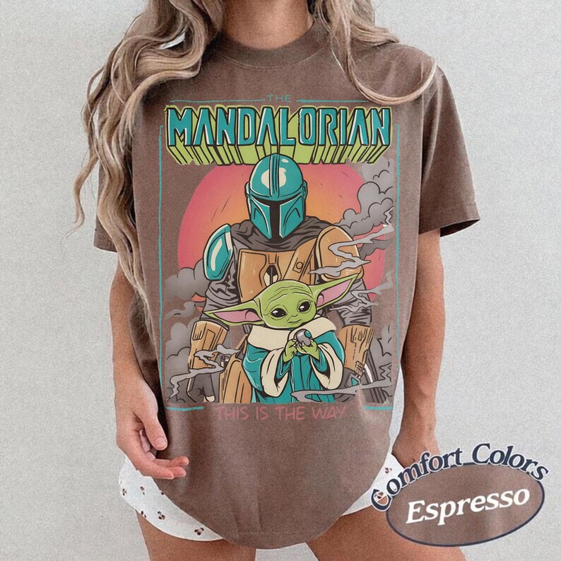 May include: A brown t-shirt featuring a graphic print of The Mandalorian and Grogu. The text THE MANDALORIAN appears at the top in teal and yellow, with THIS IS THE WAY printed at the bottom. The design includes a pink sun and grey smoke.