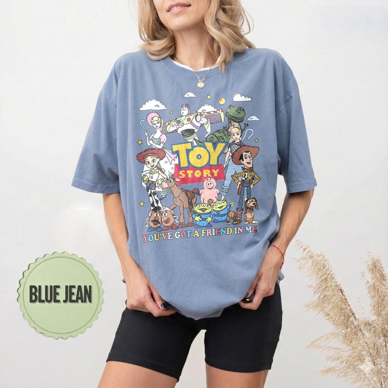 May include: A blue jean t-shirt featuring a colorful graphic of characters from the animated film "Toy Story." The shirt has the text "TOY STORY" and "YOU'VE GOT A FRIEND IN ME" printed on it. The shirt is a light blue color.
