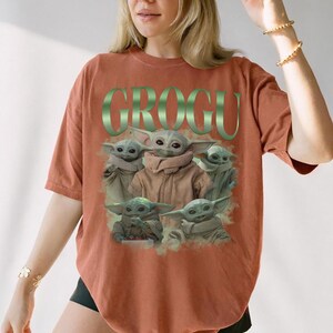 Op de afbeelding: Terracotta-kleurig T-shirt met een afbeelding van Grogu, ook bekend als Baby Yoda, uit The Mandalorian. Het woord "GROGU" is groen boven de afbeelding gedrukt. De afbeelding toont Grogu in verschillende poses.
